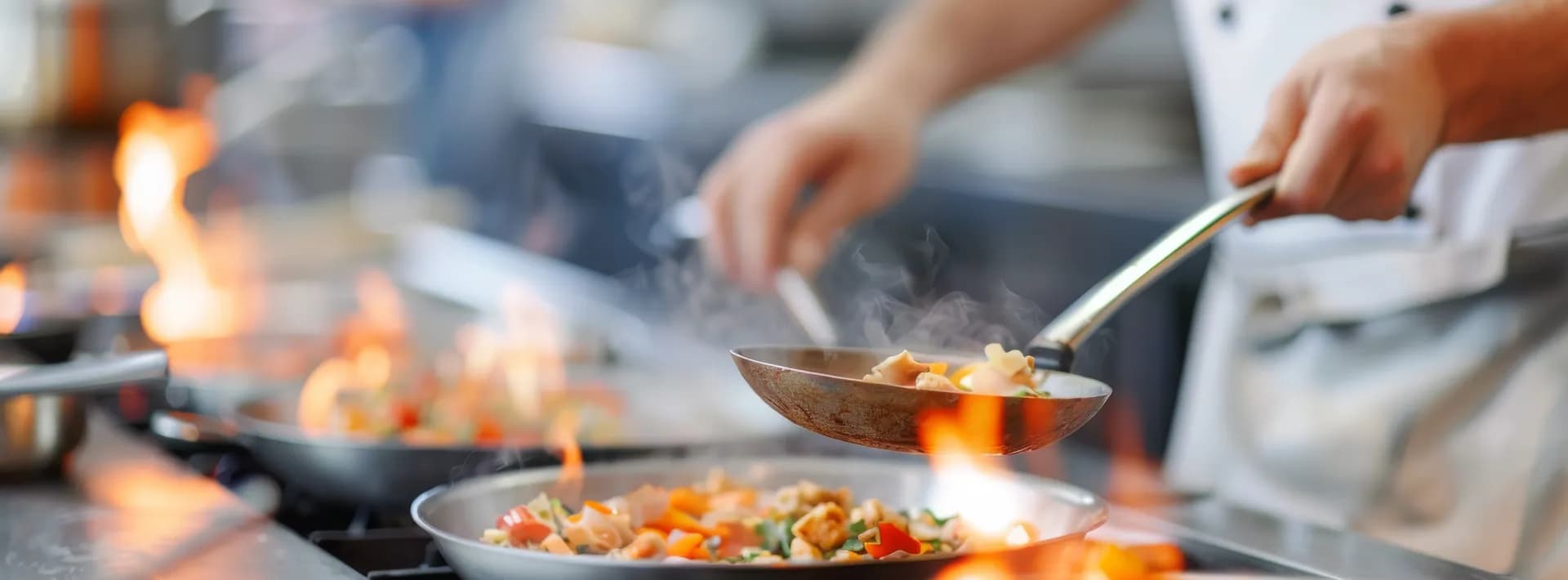 detail of sauté pans in the foreground with the arm of the chef seen working with a pan in the background