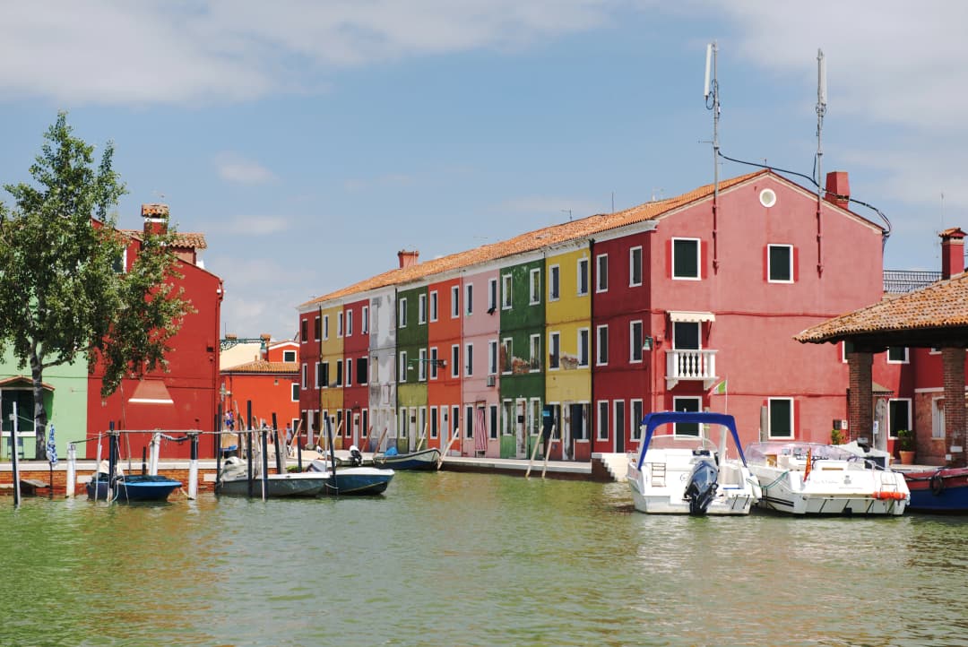View approaching a canal in Burano, Italy with anchored boats and a row colorful buildings